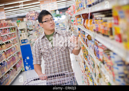Young adult Asian looks confused while reading label of cans while shopping for groceries at grocery store. Stock Photo