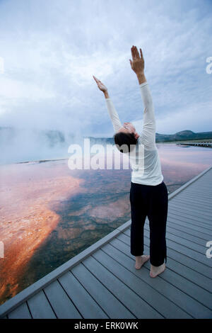 As the sun rises, a woman practices yoga on a boardwalk over algae beds while steam rises from the Grand Prismatic Spring in Yel Stock Photo