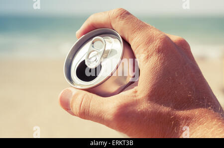 Aluminum can of beer in male hand with blurred beach and sea on a background, vintage toned photo, retro tonal photo filter corr Stock Photo
