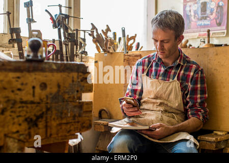 An antique furniture restorer in his workshop, using a smart phone. Stock Photo