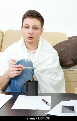 Sick man measuring temperature on thermometer while sitting on sofa at home. Ill male student  having headache covered with whit Stock Photo