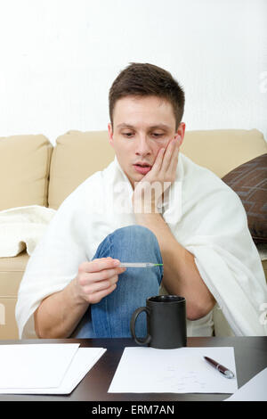 Sick man measuring temperature on thermometer while sitting on sofa at home. Ill male student  having headache covered with whit Stock Photo