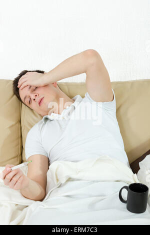 Portrait of sick man in bedroom suffering from influenza measuring temperature with thermometer and drinking tea Stock Photo