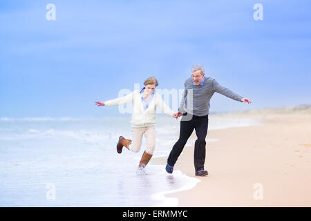 Happy middle aged couple, beautiful active woman and laughing man running on a beach jumping away from waves on the North Sea, Stock Photo