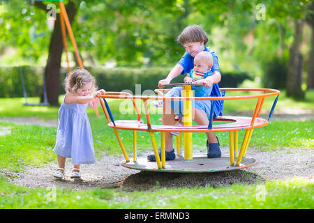 Three happy children, laughing teenager boy, cute baby and adorable toddler girl, playing together on a playground swing Stock Photo