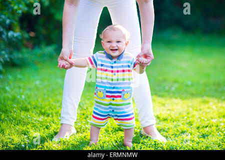 Cute funny happy baby in a colorful shirt making his first steps on green lawn in sunny summer garden, mother holding his hands Stock Photo