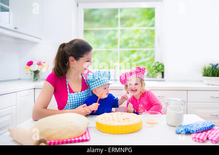Young happy mother and her kids wearing pink and blue chef hats baking a pie together in a white sunny kitchen with big window Stock Photo