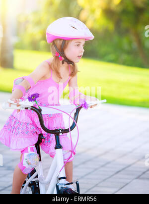 Cute little girl riding a bike in the park in bright sunny day, wearing nice pink dress, enjoying summer sport, happy childhood Stock Photo