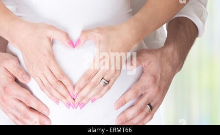 Pregnant woman and her husband holding hands on tummy in heart shape, young loving family, new life concept background Stock Photo