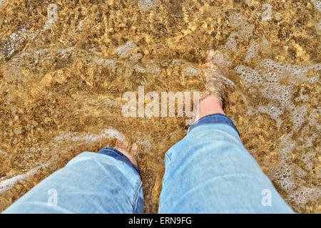 Legs of woman covered by sea wave with white sea foam in sunny day on the beach Stock Photo