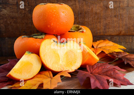 Stack of fresh Persimmons with cut slices on Autumn Fall leaves against a dark wood vintage background.
