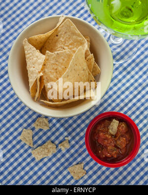 A bowl of corn tortilla chips sit beside a red bowl of salsa on a blue and white checkered tablecloth.  Pieces of tortilla are b Stock Photo