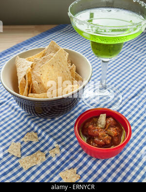 A bowl of corn tortilla chips sit beside a red bowl of salsa on a blue and white checkered tablecloth.  Pieces of tortilla are b Stock Photo