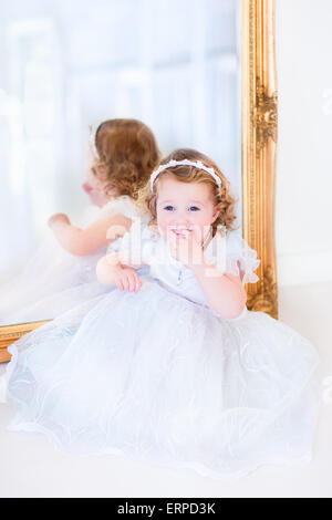 Sweet curly little girl with big beautiful eyes wearing a white bridesmaid dress sitting at a big window playing princess Stock Photo