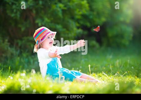 Happy laughing little girl wearing a blue dress and colorful straw hat playing with a flying butterfly having fun in the garden Stock Photo