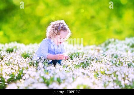 Happy laughing toddler girl with curly hair in blue dress playing with first spring snowdrops flowers in a beautiful sunny park Stock Photo