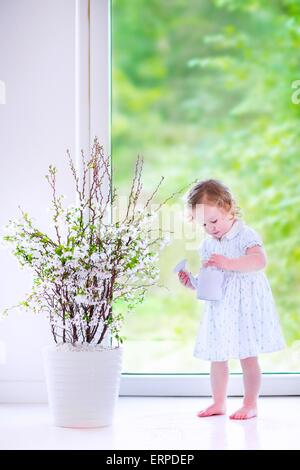 Cute little girl, funny toddler with curly hair wearing a blue festive dress, watering flowers - cherry blossom tree Stock Photo