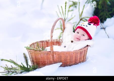 Funny happy baby boy wearing a warm snow suit and a red knitted snowman hat laying in a basket as a Christmas present in a park Stock Photo