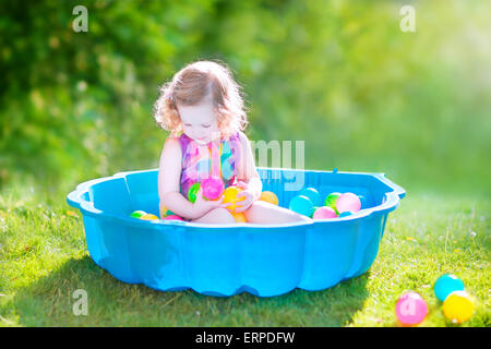 Happy cute toddler girl with curly hair wearing a pink colorful dress playing in a sand box with plastic toy balls in garden Stock Photo