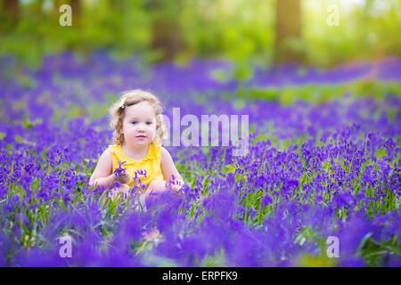 Adorable toddler girl with curly hair wearing a yellow dress playing with purple bluebell flowers in a sunny spring forest Stock Photo