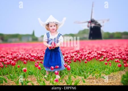 Toddler girl wearing Dutch traditional national costume dress and hat playing in a field of blooming tulips next to a windmill Stock Photo
