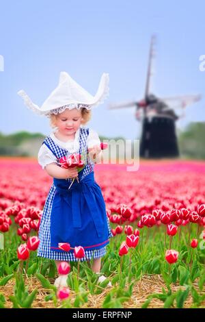 Toddler girl wearing Dutch traditional national costume dress and hat playing in a field of blooming tulips next to a windmill Stock Photo