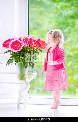 Cute happy toddler girl with curly hair wearing a pink dress playing with a bunch of beautiful big peony flowers in a vase Stock Photo