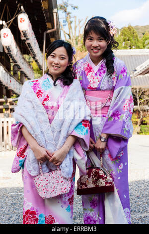 Two young Chinese women standing posing and smiling at viewer at a Japanese shrine in Kyoto. Both wearing pink and purple kimono with flower designs. Stock Photo