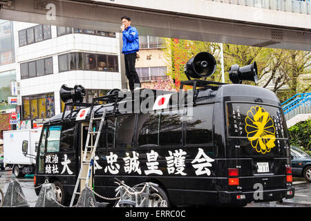 Japan, Tokyo, Harajuku. Right wing black van with political speaker holding microphone while standing on van roof haranguing people in the rain. Stock Photo