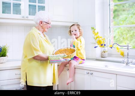 Happy beautiful great grandmother and her adorable granddaughter, curly toddler girl in colorful dress, baking an apple pie Stock Photo