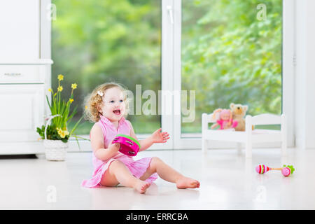 Cute curly toddler girl playing tambourine in a sunny white room with a big window Stock Photo