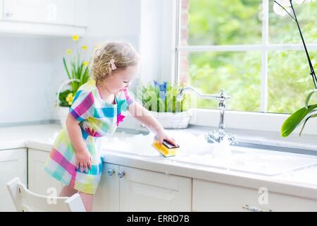 Cute curly toddler girl in a colorful dress washing dishes, cleaning with a sponge and playing with foam in the sink in kitchen Stock Photo