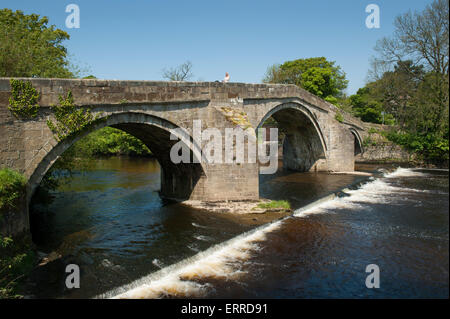 Woman crossing scenic River Wharfe on old stone packhorse bridge over flowing water & small weir - Old Bridge, Ilkley, West Yorkshire, England, UK. Stock Photo