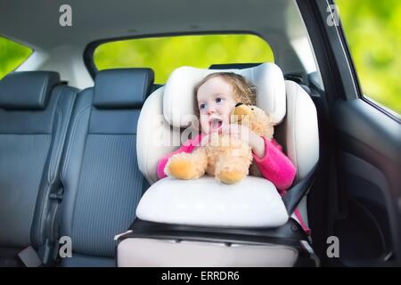 Cute curly laughing and talking toddler girl playing with a toy bear enjoying a family vacation car ride in modern safe vehicle Stock Photo