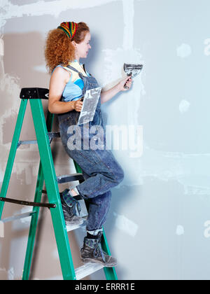 Portrait of a woman construction worker with a trowel and putty knife patching up drywall standing on a ladder Stock Photo