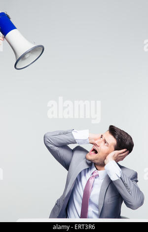 Businessman sitting at the table and covering his ears from megaphone over gray background Stock Photo
