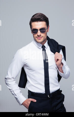 Fashion businessman with jacket on shoulder and sunglasses standing over gray background and looking at camera Stock Photo