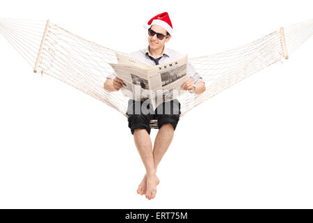 Young guy with Santa hat sitting in a hammock and reading a newspaper isolated on white background Stock Photo