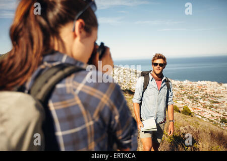 Young man posing in nature, with woman talking his pictures in countryside on a summer day while hiking. Young woman taking phot Young man posing in nature, with woman talking his pictures in countryside on a summer day while hiking. Young woman taking phot