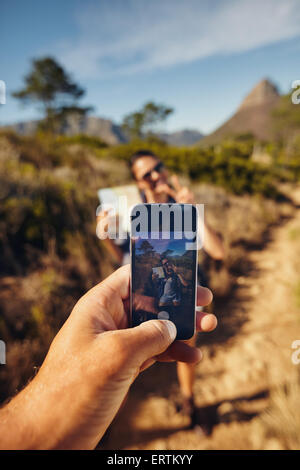 Man taking pictures on a young woman with his smartphone. Focus on mobile phone in hand. Couple hiking in countryside. Stock Photo