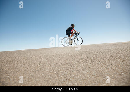Low angle image of a male cyclist riding on a flat road against blue sky. Man cycling up hill on open road. Stock Photo