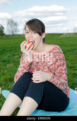 young woman sitting outdoors on a blanket and eating an apple Stock Photo