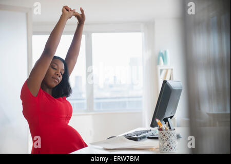 Black pregnant businesswoman stretching arms at desk in office Stock Photo