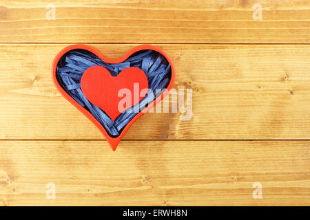 Red painted wooden heart enclosed with blue paper raffia strips in red box on wooden background Stock Photo