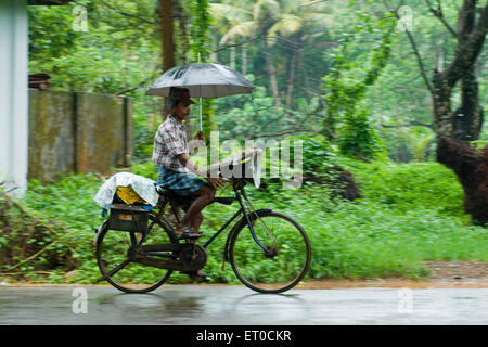 Lottery ticket seller riding bicycle with umbrella in rain ; Kerala ; India Stock Photo