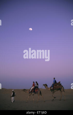 Tourist sitting on camels fall of night with moon at Sam sand dunes ; Jaisalmer ; Rajasthan ; India