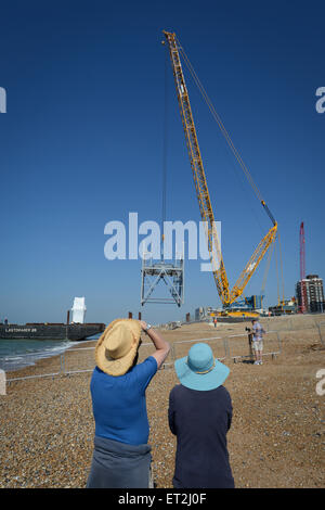 During the construction of the British Airways i360 on the beach in Brighton, East Sussex, England a man and women watch as a large crane removes sections of the tower from a barge on the beach. Stock Photo
