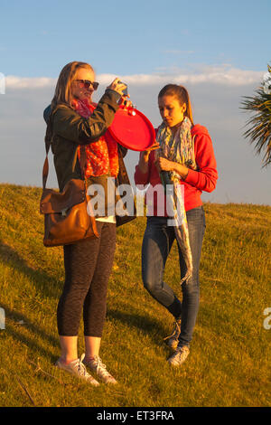 Two young women standing on grass bank with one taking a photo of the view at Sandbanks, Poole, Dorset, UK in June Stock Photo
