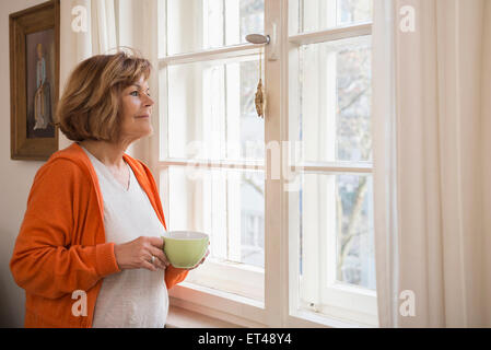 Side profile of a senior woman having cup of tea looking through window at home, Munich, Bavaria, Germany