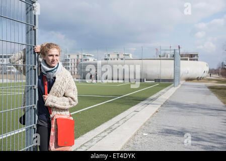 Young woman with metal fence at soccer field with players in the background Munich Bavaria Germany
